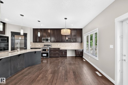 Kitchen featuring wood-finish flooring, dark wood cabinetry, stainless steel appliances, a stone countertop island, and pendant lighting - 3329 West Court Nw, Edmonton, AB 