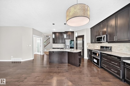 Open-concept kitchen with dark wood cabinetry, stainless steel appliances, light-toned countertops, and a central island with pendant lighting - 3329 West Court Nw, Edmonton, AB 
