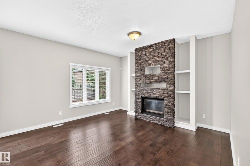 Stacked stone fireplace with built-in shelving, wood-finish flooring, and natural light from a large window - 3329 West Court Nw, Edmonton, AB 