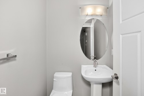 Pedestal sink with chrome faucet, oval mirror, wall-mounted light fixture, and white toilet - 3329 West Court Nw, Edmonton, AB 