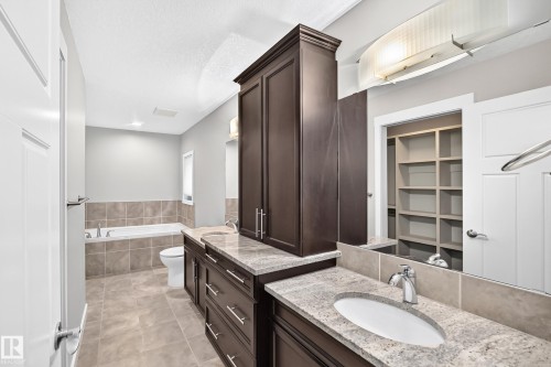 Dual vanity with stone countertops, dark wood cabinetry, a built-in soaking tub with tile surround, and a walk-in closet with shelving - 3329 West Court Nw, Edmonton, AB 