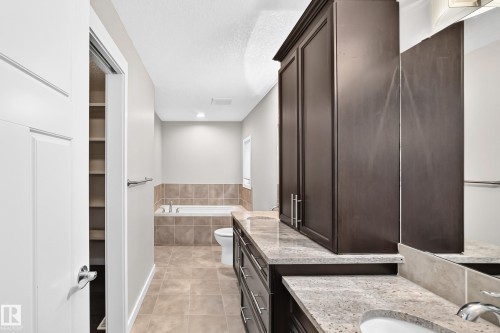 Bathroom featuring a built-in tub with tile surround, tile flooring, dark wood-finish cabinetry, light-toned stone countertops, and a recessed storage area with shelving - 3329 West Court Nw, Edmonton, AB 
