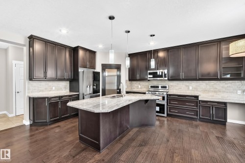 Spacious kitchen featuring dark wood-finish cabinetry, light-toned countertops, stainless steel appliances, a large center island with a sink, and wood-finish flooring - 3329 West Court Nw, Edmonton, AB 
