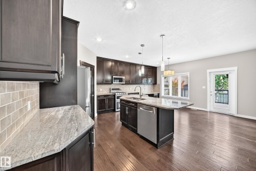 Kitchen featuring dark wood-finish cabinetry, light-toned countertops, subway tile backsplash, stainless steel appliances, and a central island with an undermount sink - 3329 West Court Nw, Edmonton, AB 