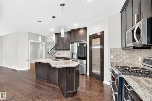 Kitchen featuring wood-finish flooring, dark wood cabinetry, stainless steel appliances, a tile backsplash, and an island with a light-toned countertop and pendant lighting - 3329 West Court Nw, Edmonton, AB 