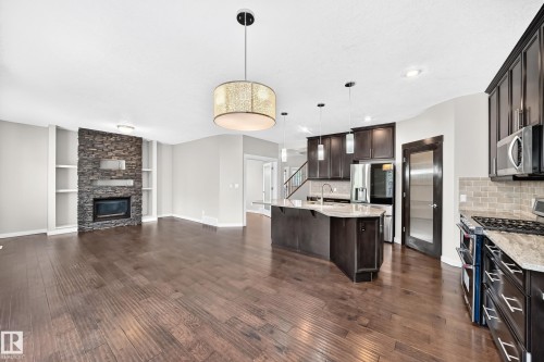 Dark wood-finish flooring, a stacked stone fireplace with built-in shelving, dark wood cabinetry, a kitchen island with stone countertops, and stainless steel appliances - 3329 West Court Nw, Edmonton, AB 
