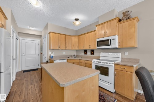 Kitchen featuring wood-finish flooring, a central island with a solid surface countertop, wood cabinetry, and white appliances including a microwave and range - #420 9820 165 St Nw, Edmonton, AB 