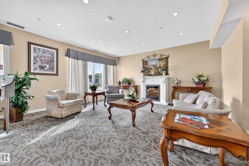 Recessed lighting illuminates the neutral-toned walls, decorative fireplace with a mantel, patterned carpeting, and sheer window treatments - #420 9820 165 St Nw, Edmonton, AB 