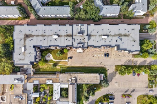 Aerial perspective highlighting a large building with a flat roof, an interior courtyard featuring a circular landscaped area, adjacent parking areas, and surrounding mature trees - #420 9820 165 St Nw, Edmonton, AB 
