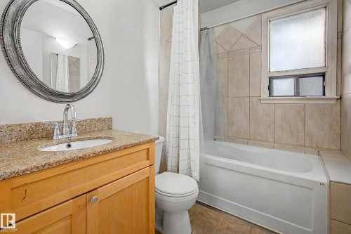 Bathroom featuring a light wood-finish vanity, granite countertop with an undermount sink, a framed oval mirror, tiled tub surround, and a built-in bathtub - 10830 138 Street Nw, Edmonton, AB 
