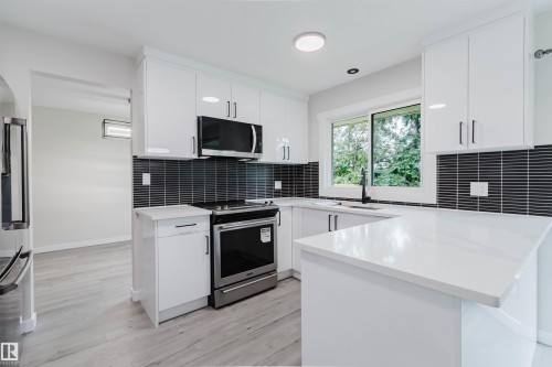 Modern kitchen featuring white cabinetry, light-toned wood-finish flooring, stainless steel appliances, dark tile backsplash, and a corner sink with a window - 10423 35 Avenue Nw, Edmonton, AB 