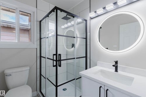 Modern bathroom featuring a black-framed glass shower enclosure, white vanity with integrated sink, illuminated circular mirror, and matte black fixtures - 10423 35 Avenue Nw, Edmonton, AB 
