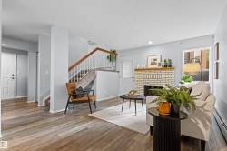 Wood-finish flooring throughout, featuring a white brick fireplace with a wood mantle, a staircase with white spindles and wood handrail, and white panel doors - 