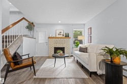 Light-colored brick fireplace with hearth, wood-finish flooring, staircase with carpeted treads and wood handrail, and white paneled door with glass insert - 