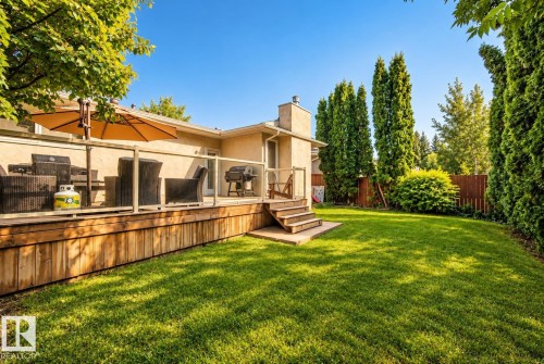 Expansive backyard featuring a wood deck with glass panel railings, a natural stone chimney, mature evergreen trees, and a privacy fence - 11 Brunswick Crescent, St. Albert, AB 