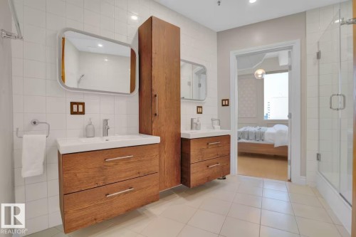 Dual floating wood-finish vanities with vessel sinks, a tall wood-finish storage cabinet, white square tiled walls, and a glass-enclosed shower - #904 10105 109 St Nw, Edmonton, AB 