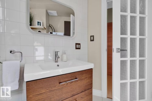 White subway tile wall, rounded rectangular mirror, integrated sink, wood-finish vanity with drawer pulls, and a frosted paneled door - #904 10105 109 St Nw, Edmonton, AB 