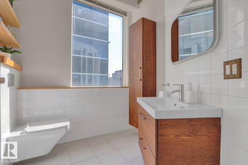 Modern bathroom featuring a wall-mounted toilet, wood-finish vanity with integrated sink, and white tiled walls - #904 10105 109 St Nw, Edmonton, AB 