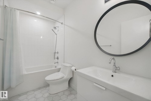 White bathroom featuring a built-in tub with tile surround, hexagonal floor tiles, a vanity with an integrated sink, a chrome faucet, and a round wall mirror - 10925 University Avenue Nw, Edmonton, AB 