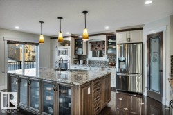 Spacious kitchen featuring a large island with a stone-finish countertop, three pendant lights, stainless steel appliances, and a glass-paneled pantry door - 
