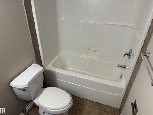 White toilet, white bathtub with integrated shower, chrome fixtures, and neutral-toned wall tiles - #205 10335 117 St Nw, Edmonton, AB 