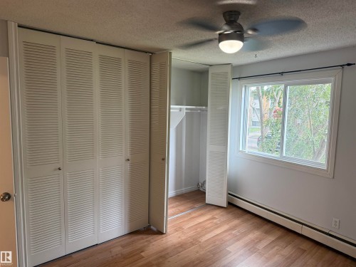 Wood-finish flooring, a ceiling fan with integrated lighting, louvered bi-fold closet doors, a built-in closet rod and shelving, and a large window with white trim - #205 10335 117 St Nw, Edmonton, AB 