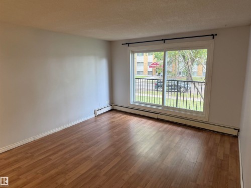 Wood-finish flooring, white painted walls, and a sliding glass door to an exterior balcony with black metal railing - #205 10335 117 St Nw, Edmonton, AB 