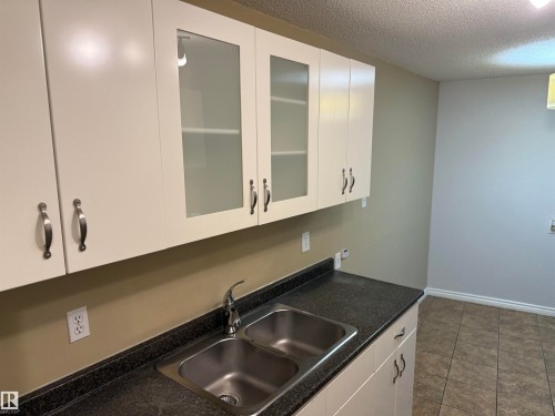 White cabinetry with brushed nickel hardware, a dual basin stainless steel sink, black speckled countertops, and a tiled floor - #205 10335 117 St Nw, Edmonton, AB 