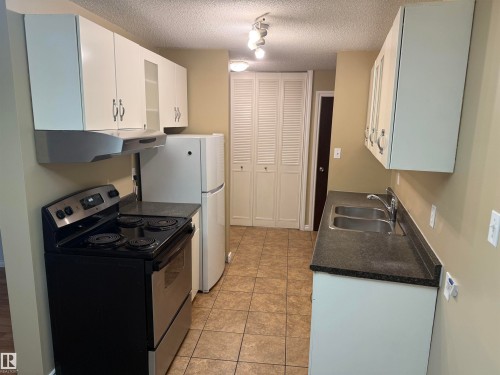 Kitchen featuring white cabinetry, dark countertops, a double basin stainless steel sink, and a range with a coordinating hood - #205 10335 117 St Nw, Edmonton, AB 