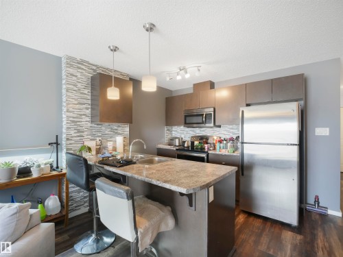 Kitchen featuring stainless steel appliances, wood-finish flooring, a stone-finish countertop island, modern pendant lighting, and a textured tile backsplash - #414 340 Windermere Rd Nw, Edmonton, AB 