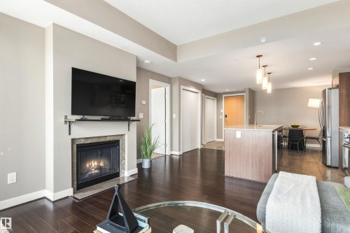 Open-concept living area featuring a fireplace with stone surround, wood-finish flooring, a kitchen island with built-in sink, and pendant lighting - #416 2510 109 St Nw, Edmonton, AB 