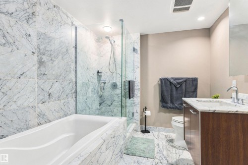 Porcelain bathtub with surrounding natural stone tile, glass-enclosed shower, wood-finish vanity with stone countertop, and chrome fixtures - #416 2510 109 St Nw, Edmonton, AB 