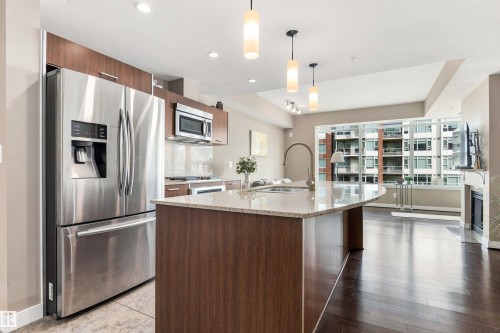 Stainless steel appliances, wood-finish cabinetry, curved kitchen island with integrated sink, recessed lighting, and dark wood-finish flooring - #416 2510 109 St Nw, Edmonton, AB 