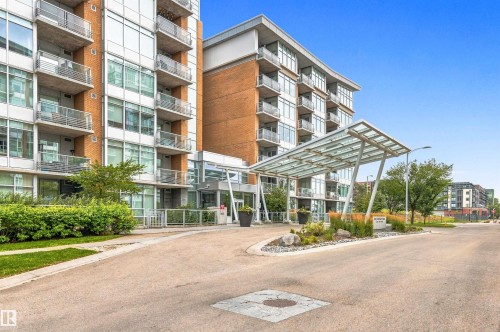Modern high-rise building with brick and glass facade, featuring multiple balconies with metal railings, a covered entryway with a glass canopy, and landscaping with shrubs and natural stone elements - #416 2510 109 St Nw, Edmonton, AB 