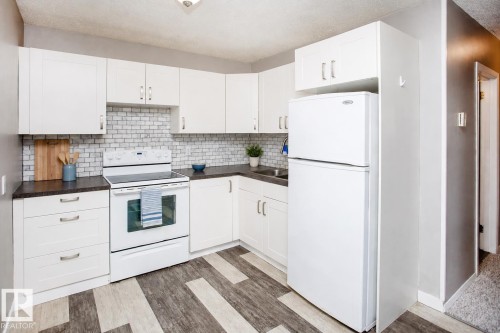 Kitchen featuring white cabinetry, subway tile backsplash, dark countertops, white appliances, and multi-tone wood-finish flooring. VIRTUALLY STAGED - #8 15431 93 Av Nw, Edmonton, AB 