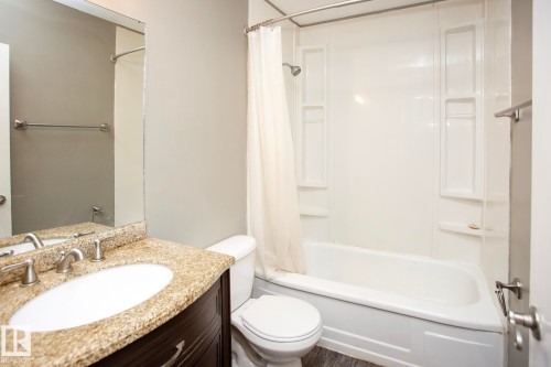 Bathroom featuring a white bathtub with integrated shelving, a built-in toilet, a vanity with a granite countertop, an undermount sink, and brushed nickel hardware - #8 15431 93 Av Nw, Edmonton, AB 