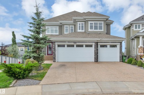 Two-story residence featuring a dual garage with stone accents, stucco exterior, mature evergreen landscaping, and an exposed aggregate driveway - 9054 24 Avenue Sw, Edmonton, AB 