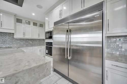 Gourmet kitchen featuring a large stainless steel refrigerator, built-in double ovens, white shaker cabinetry, an expansive island with a waterfall edge, and mosaic tile backsplash - 9054 24 Avenue Sw, Edmonton, AB 