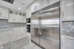 Gourmet kitchen featuring a large stainless steel refrigerator, built-in double ovens, white shaker cabinetry, an expansive island with a waterfall edge, and mosaic tile backsplash - 