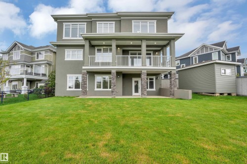 Expansive green lawn with a two-story stucco exterior, upper-level covered balcony with white railings, ground-level patio, and stone column detailing - 9054 24 Avenue Sw, Edmonton, AB 