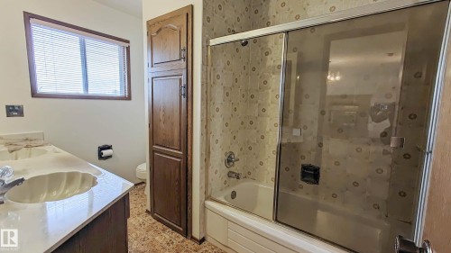 Bathroom featuring a double vanity with shell-shaped sinks, a built-in storage cabinet, a window with blinds, and a tub with a sliding glass enclosure - 3031 79 Street Nw, Edmonton, AB 