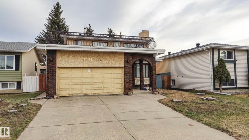 Brown brick archway entrance, attached garage, concrete driveway, upper-level balcony with metal railing, and a mature evergreen tree - 3031 79 Street Nw, Edmonton, AB 