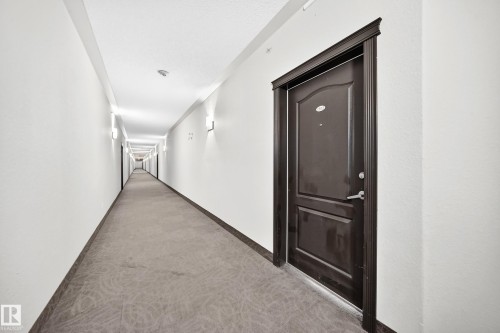 Long hallway featuring wall-mounted sconce lighting, light-toned walls, dark wood-finish door frames, and patterned carpeting - #1303 330 Clareview Station Dr Nw, Edmonton, AB 