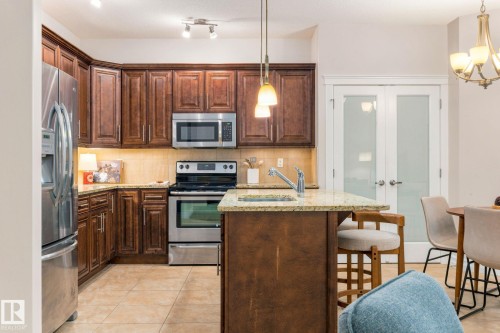 Kitchen featuring dark wood cabinetry, stainless steel appliances, light-toned tiled flooring, and a central island with a sink and pendant lighting - 10143 123 Street Nw, Edmonton, AB 