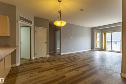 Spacious room featuring wood-finish flooring, light grey painted walls, a sliding glass door leading to an outdoor balcony, two vertical windows, and an overhead pendant light fixture - #412 4304 139 Av Nw, Edmonton, AB - Indoor Photo Showing Kitchen