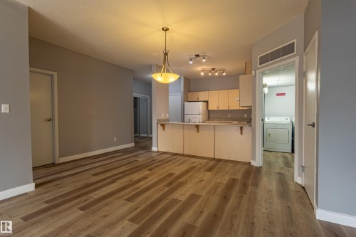 Open concept living space featuring wood-finish flooring, a kitchen with light wood cabinetry and track lighting, and a separate laundry area with a washer and dryer unit - #412 4304 139 Av Nw, Edmonton, AB - Indoor Photo Showing Kitchen