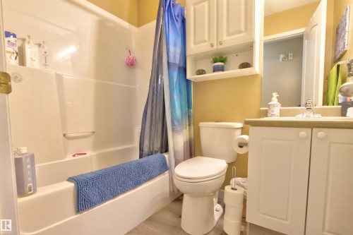 Bathroom featuring a white integrated tub and shower unit, a vanity with white cabinetry and a light-toned countertop, a toilet, an over-toilet wall cabinet with open shelving, and wood-finish flooring - #106 2 Alpine Bv, St. Albert, AB - Indoor Photo Showing Bathroom