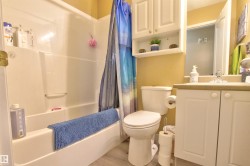 Bathroom featuring a white integrated tub and shower unit, a vanity with white cabinetry and a light-toned countertop, a toilet, an over-toilet wall cabinet with open shelving, and wood-finish flooring - 