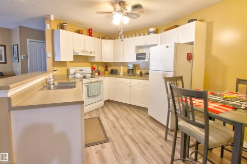 Open-concept kitchen featuring wood-finish flooring, white cabinetry, integrated appliances, and a ceiling fan - #106 2 Alpine Bv, St. Albert, AB - Indoor Photo Showing Kitchen With Double Sink