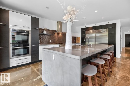 Gourmet kitchen with stainless steel double ovens, large center island with seating, a built-in stainless steel refrigerator, dark wood-finish cabinetry, and a mosaic tile backsplash - 14A Birch Drive, St. Albert, AB 